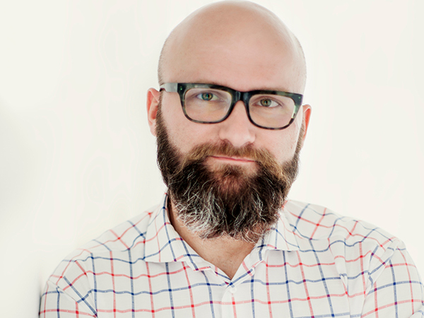 Dr. hab. Tomasz Piekot, oblečený v kostkované košili, se opírá o zeď se zkříženýma rukama.